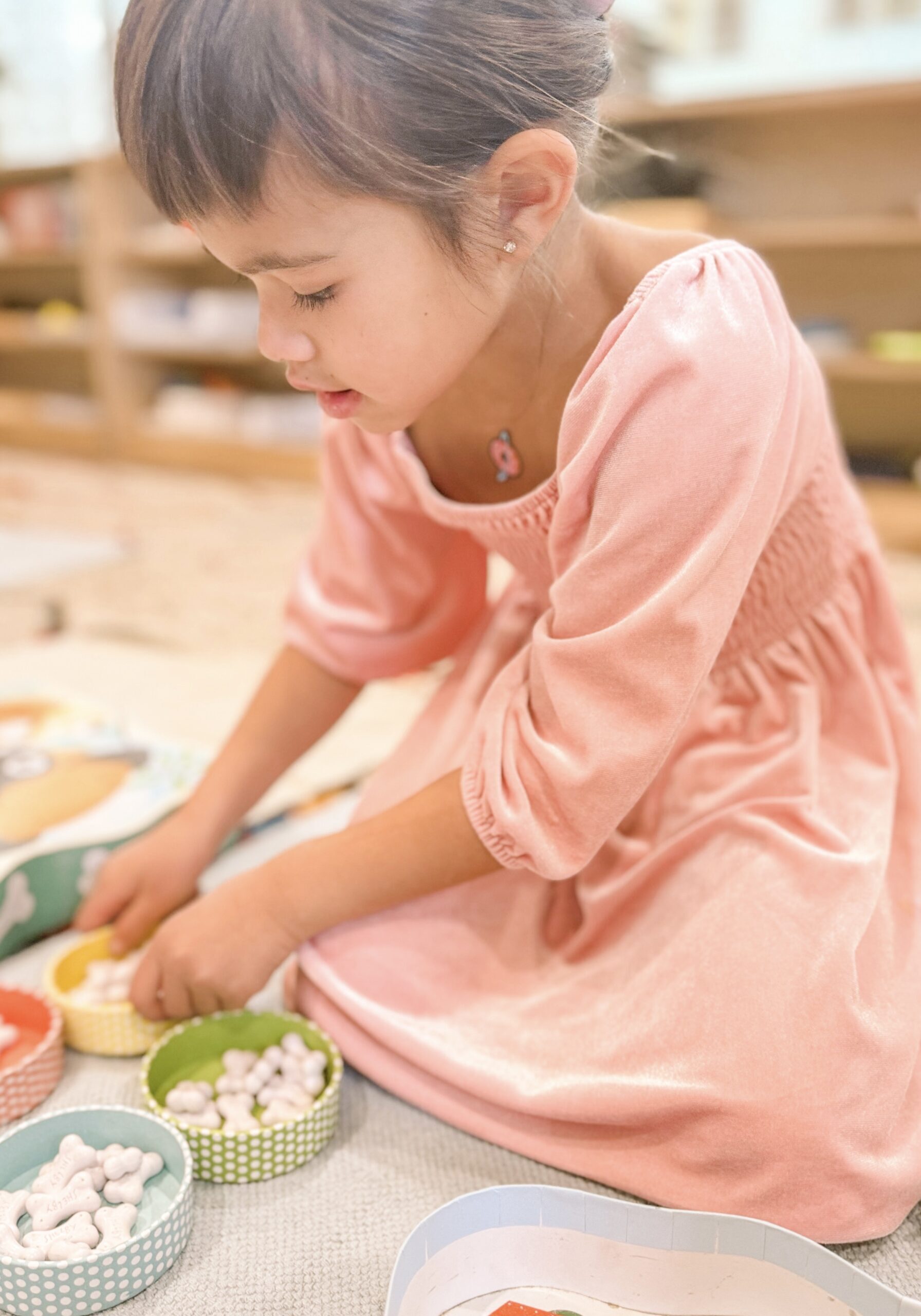 a young girl with dark hair in a pink dress playing a game sitting on the floor in a montessori preschool